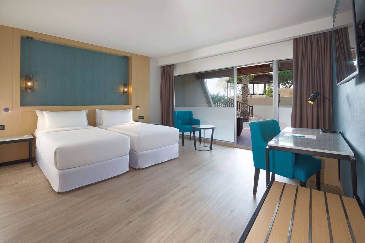 A hotel room with two beds, a desk, chairs, and a balcony with curtains.
