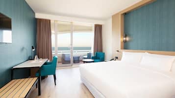 A hotel room with a large bed, a desk, a chair, and a view of the sea through a sliding door.
