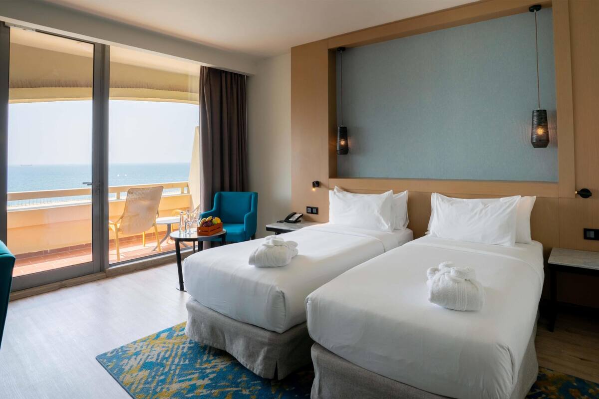 A hotel room with two beds, a balcony with a chair, a small table with a fruit bowl, and a painting on the wall.