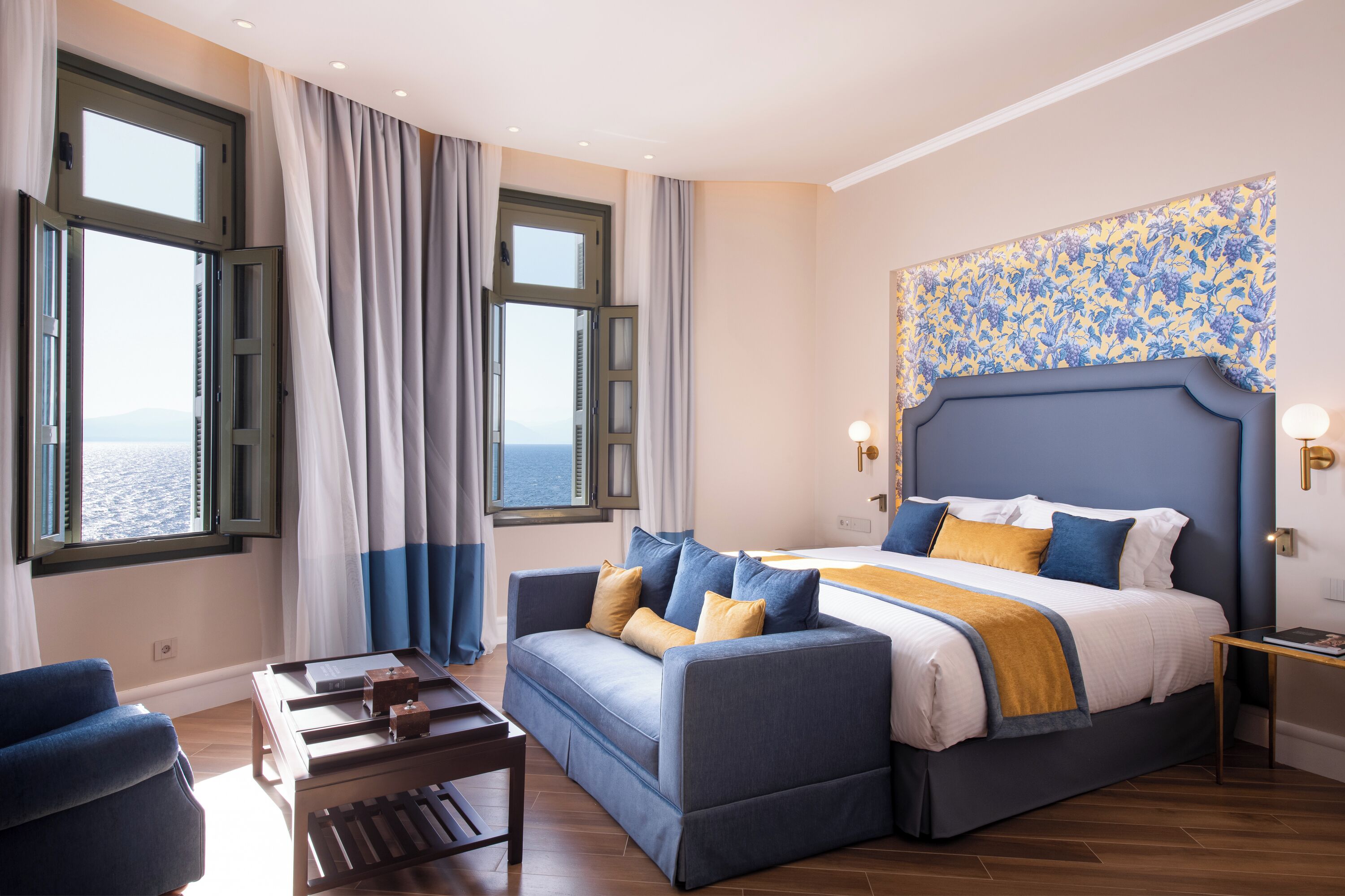 A bedroom with a bed, a sofa, a table, and a view of the sea through large windows.