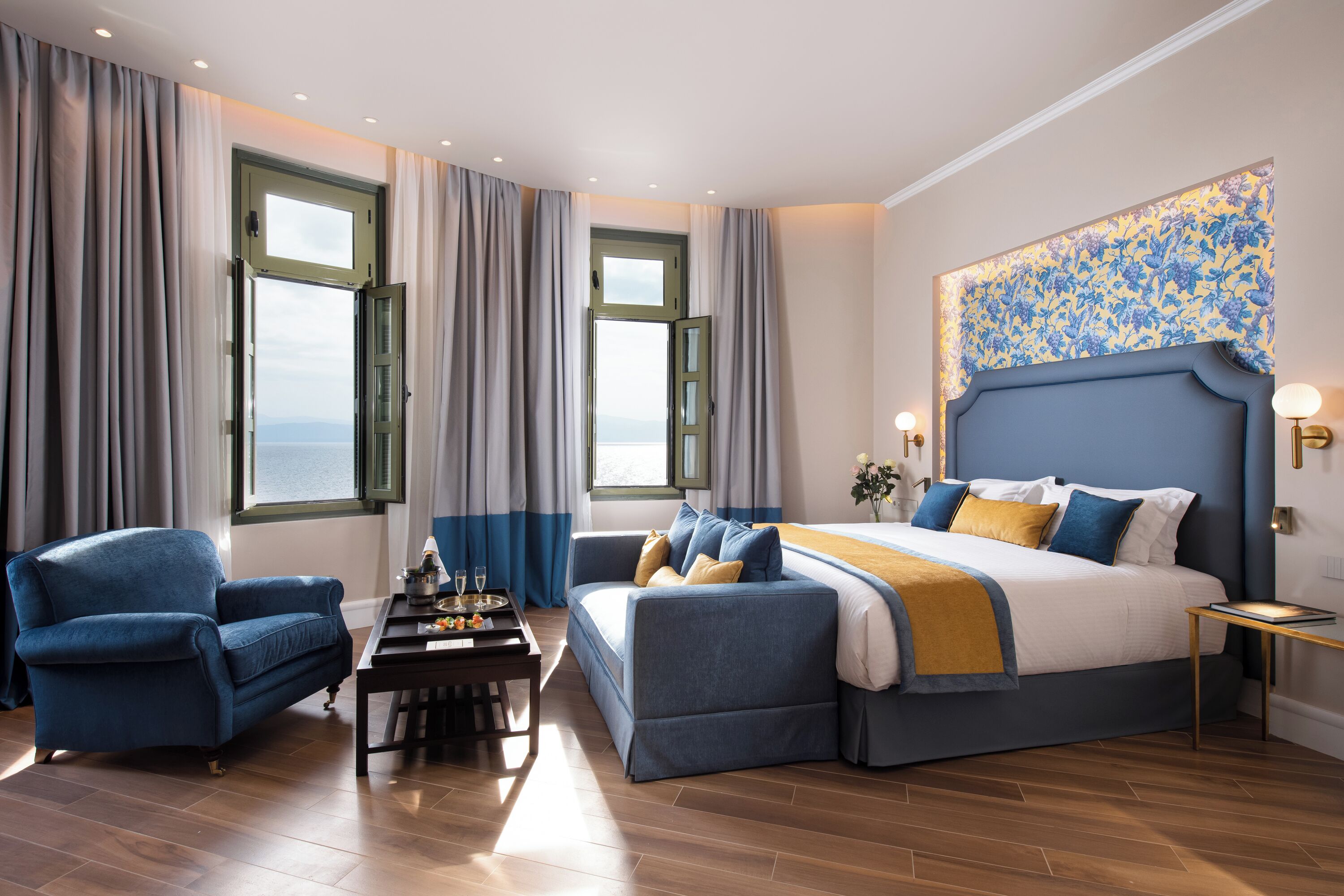 Ein Schlafzimmer mit einem großen Bett, zwei Sesseln, einem kleinen Tisch und Meerblick durch Fenster.