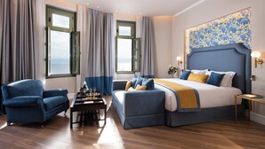A bedroom with a large bed, two armchairs, a small table, and a view of the sea through windows.