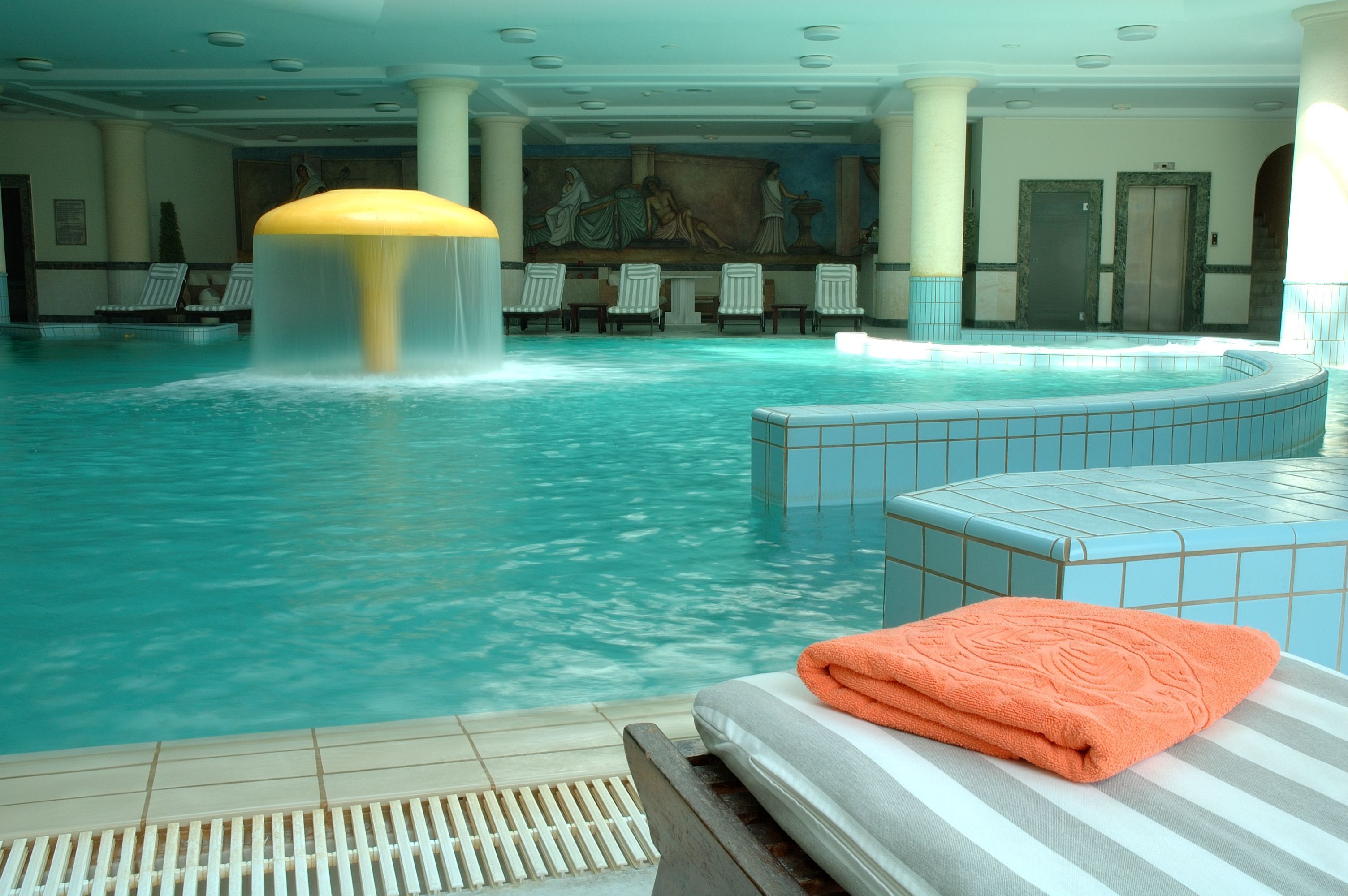 A large indoor swimming pool with a water fountain, lounge chairs, and a tiled area with a towel.