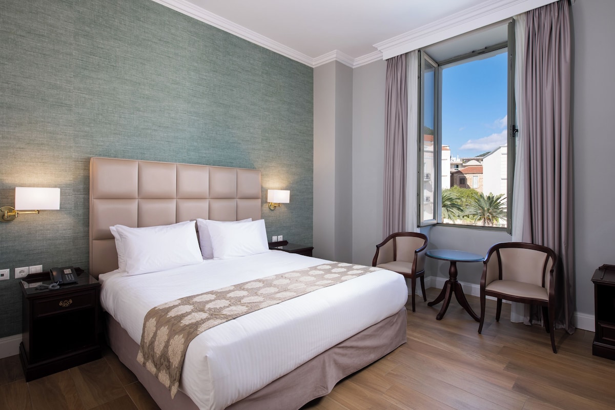 A hotel room with a large bed, bedside lamps, a small table with chairs, and a window with curtains.