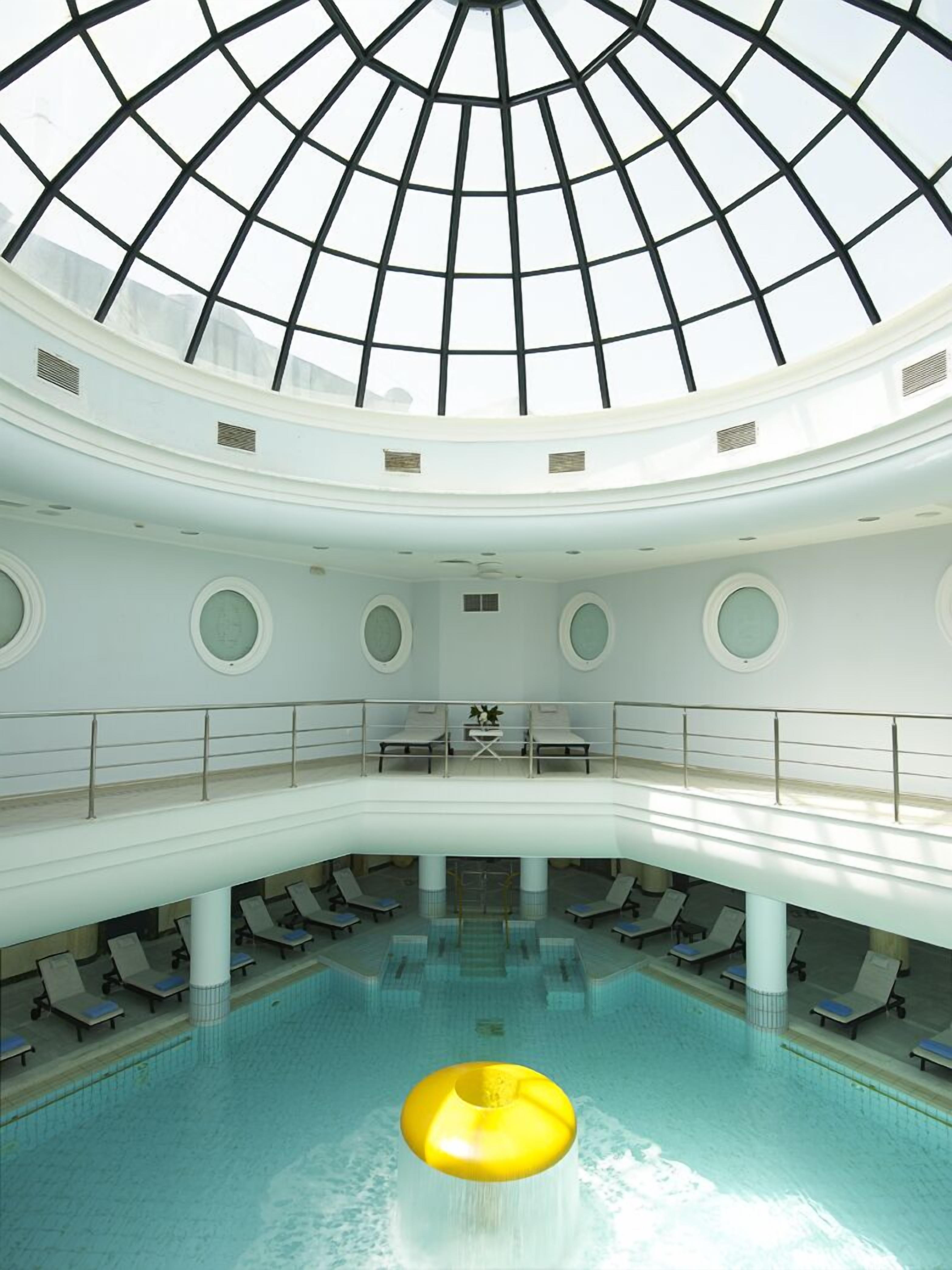 A multi-level indoor swimming pool with a glass ceiling, multiple seating areas, and a central water feature.