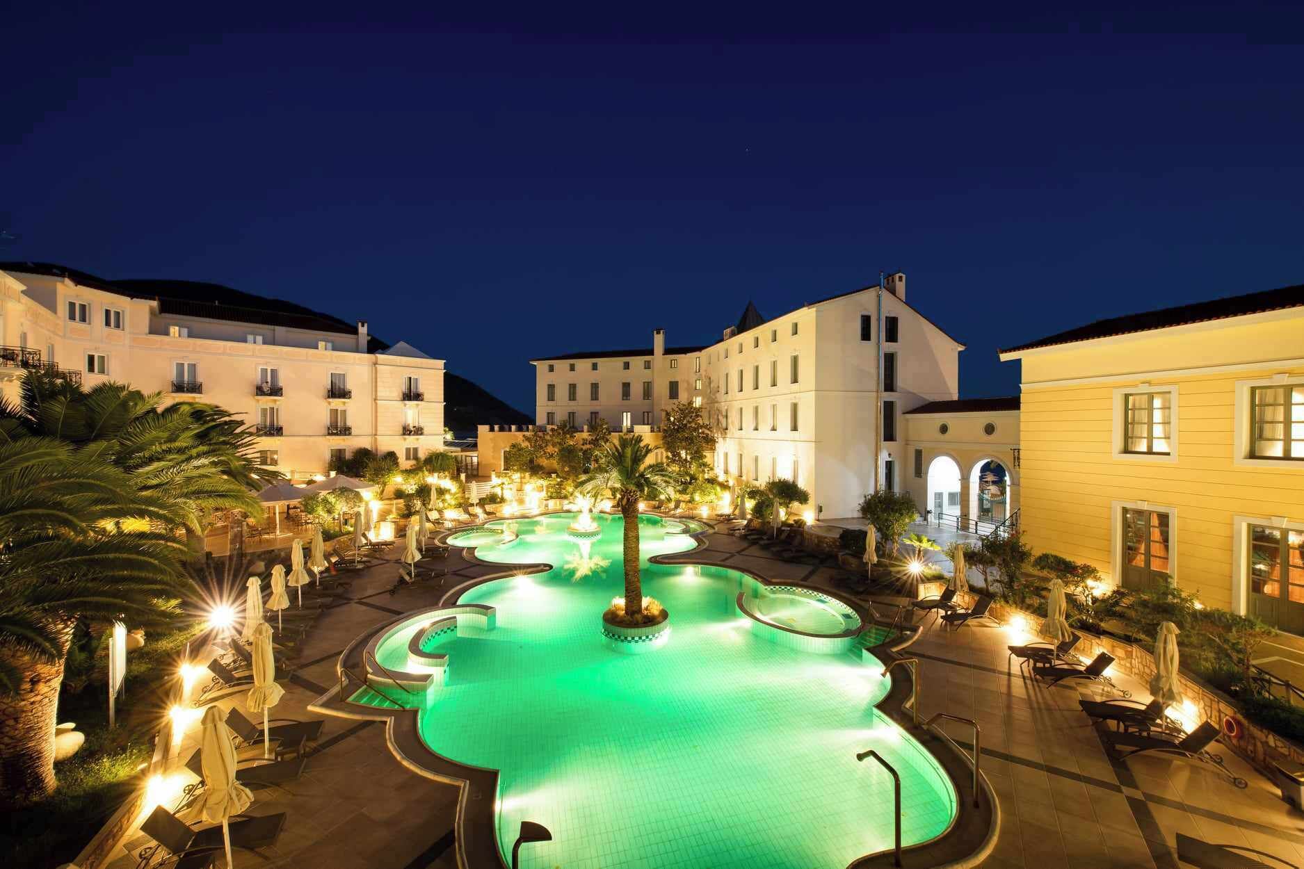 A nighttime view of a hotel courtyard with a swimming pool, palm trees, and outdoor seating areas.