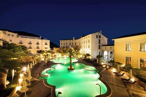 Vue nocturne d’une cour d’hôtel avec une piscine, des palmiers et des espaces de détente en plein air.