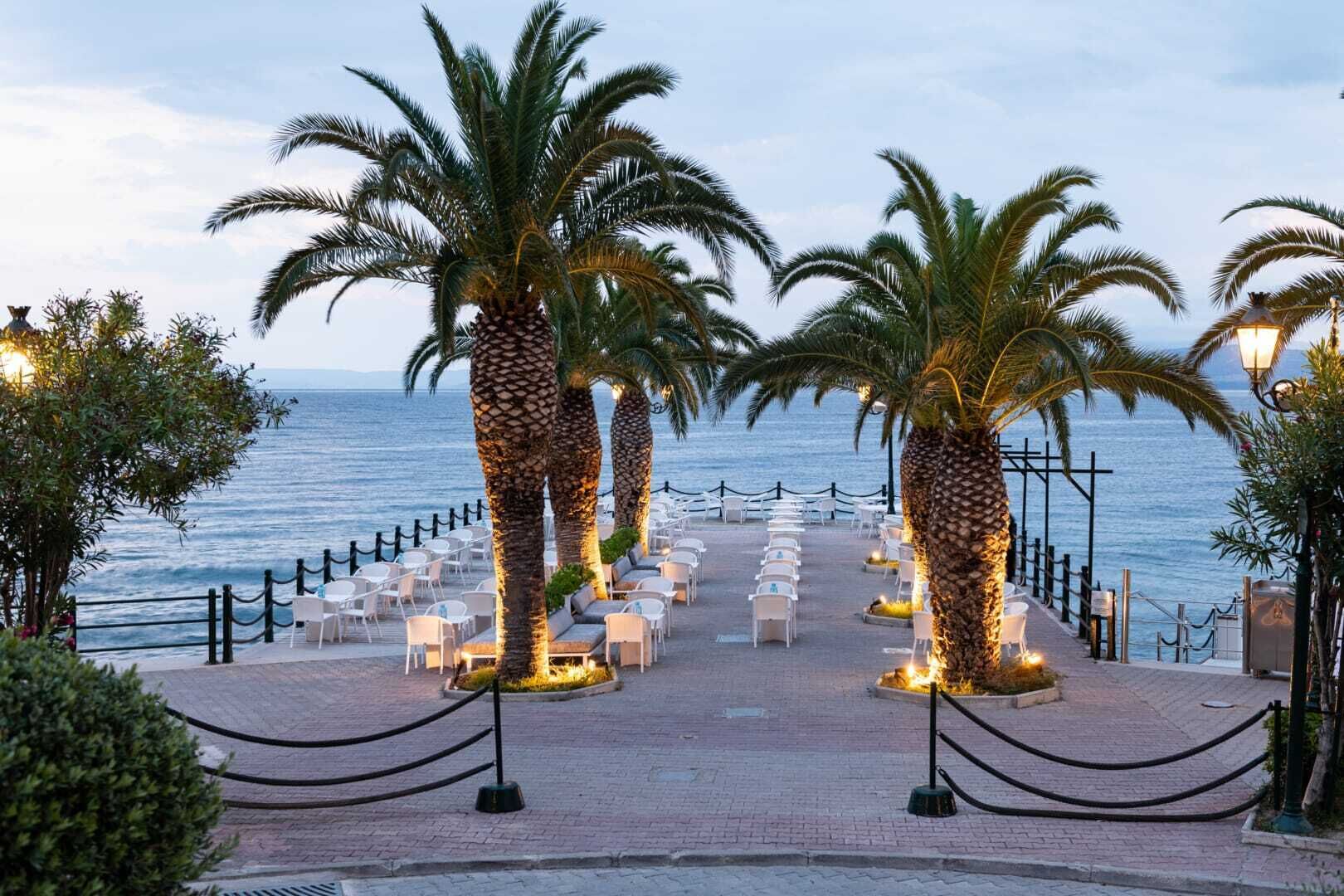 Une promenade en bord de mer, bordée de palmiers, avec des chaises longues blanches et une vue sur l’océan.