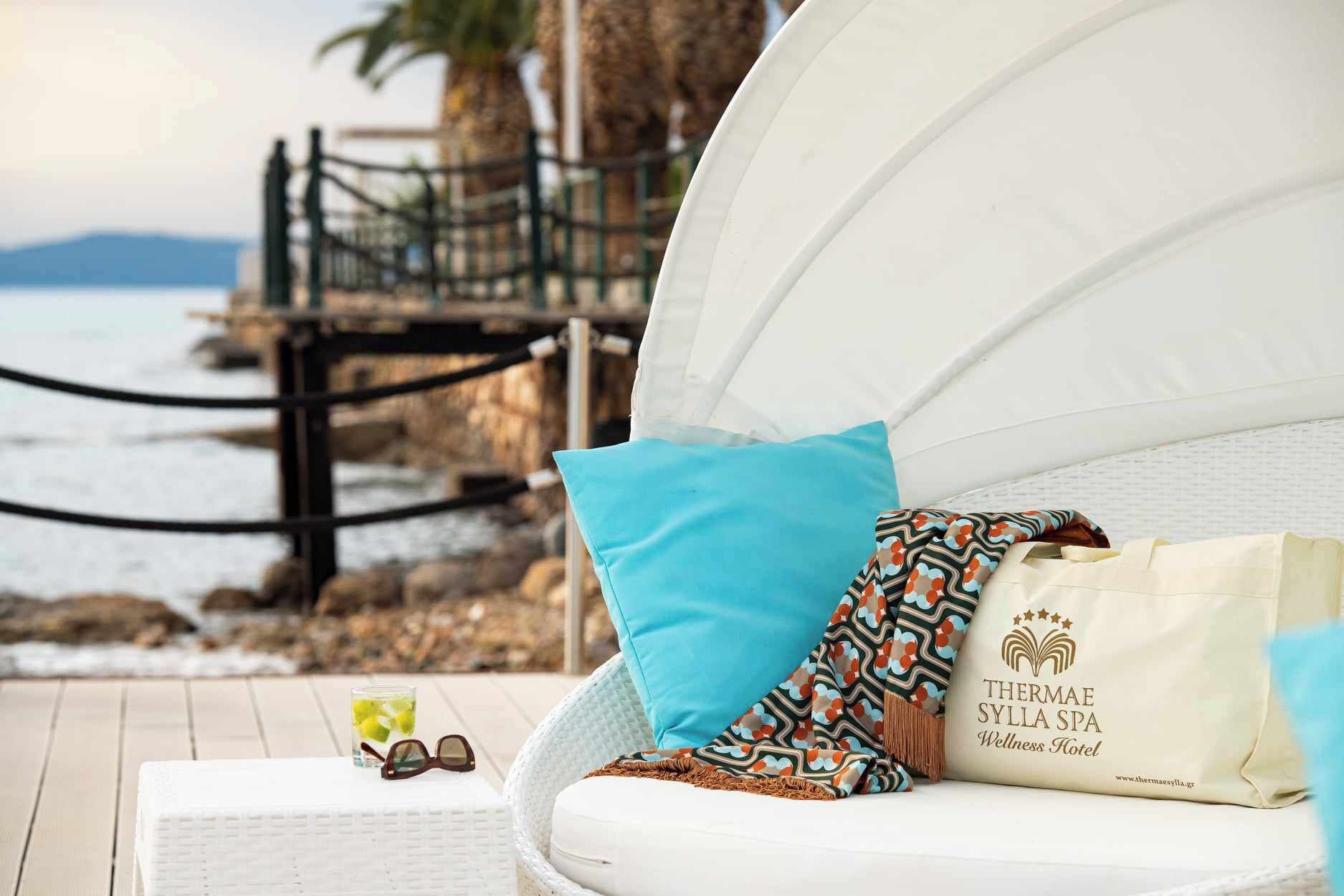 Sillón lounge blanco con cojín azul y manta estampada, un vaso de bebida, y un bolso con el logotipo del Spa Thermae Syllia sobre una terraza de madera con vistas a la costa rocosa.