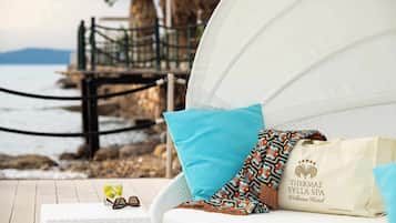 Sillón lounge blanco con cojín azul y manta estampada, un vaso de bebida, y un bolso con el logotipo del Spa Thermae Syllia sobre una terraza de madera con vistas a la costa rocosa.