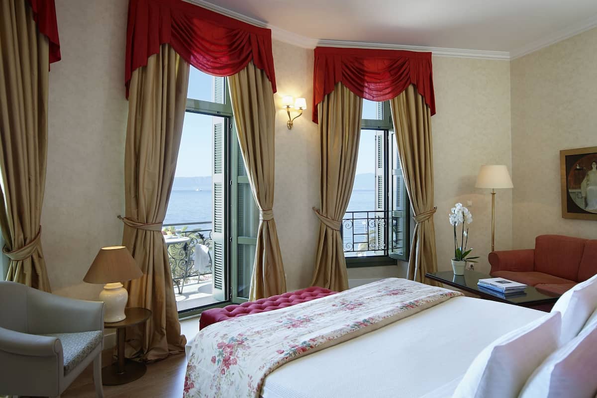 Una habitación de hotel con una cama, una silla, un escritorio, una lámpara, un jarrón con flores y un cuadro en la pared.