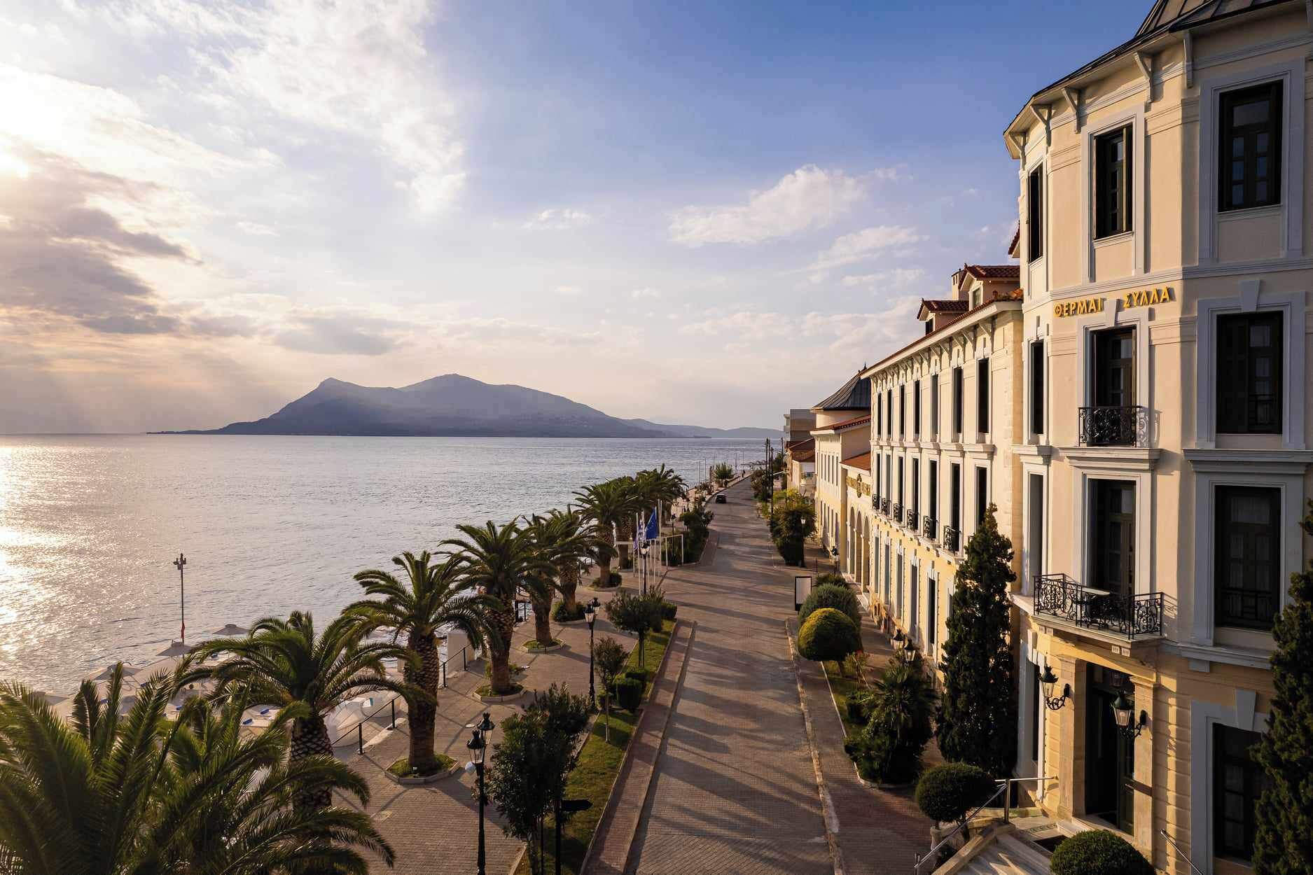 A coastal promenade with buildings, palm trees, and a view of the sea and mountains.