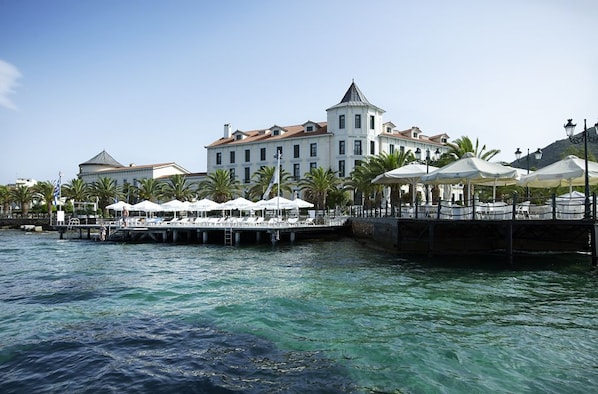 Un hotel frente al mar con un gran edificio, un muelle con sombrillas y palmeras.