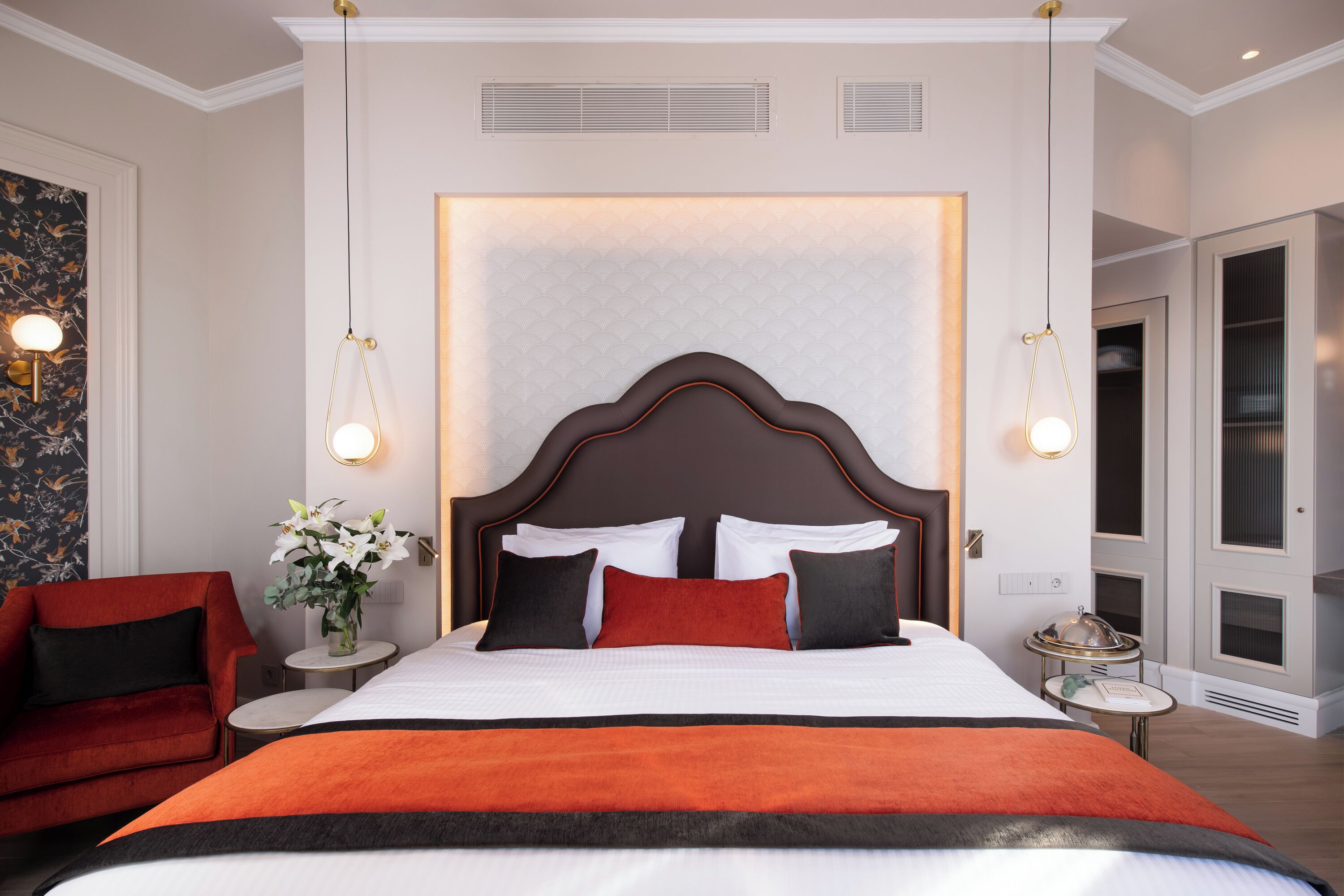 Ein Hotelzimmer mit einem großen Bett, einem roten Sessel, einem kleinen Tisch mit Blumenvase und Blick auf eine Tür.