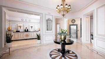A hotel lobby with a reception desk, a chandelier, a clock, and a round table with a decorative floor design.