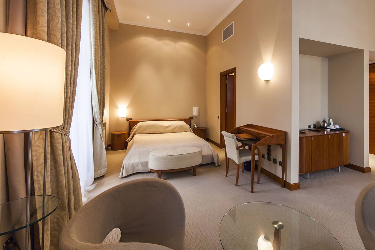 A hotel room with a bed, desk, chairs, and a lamp.