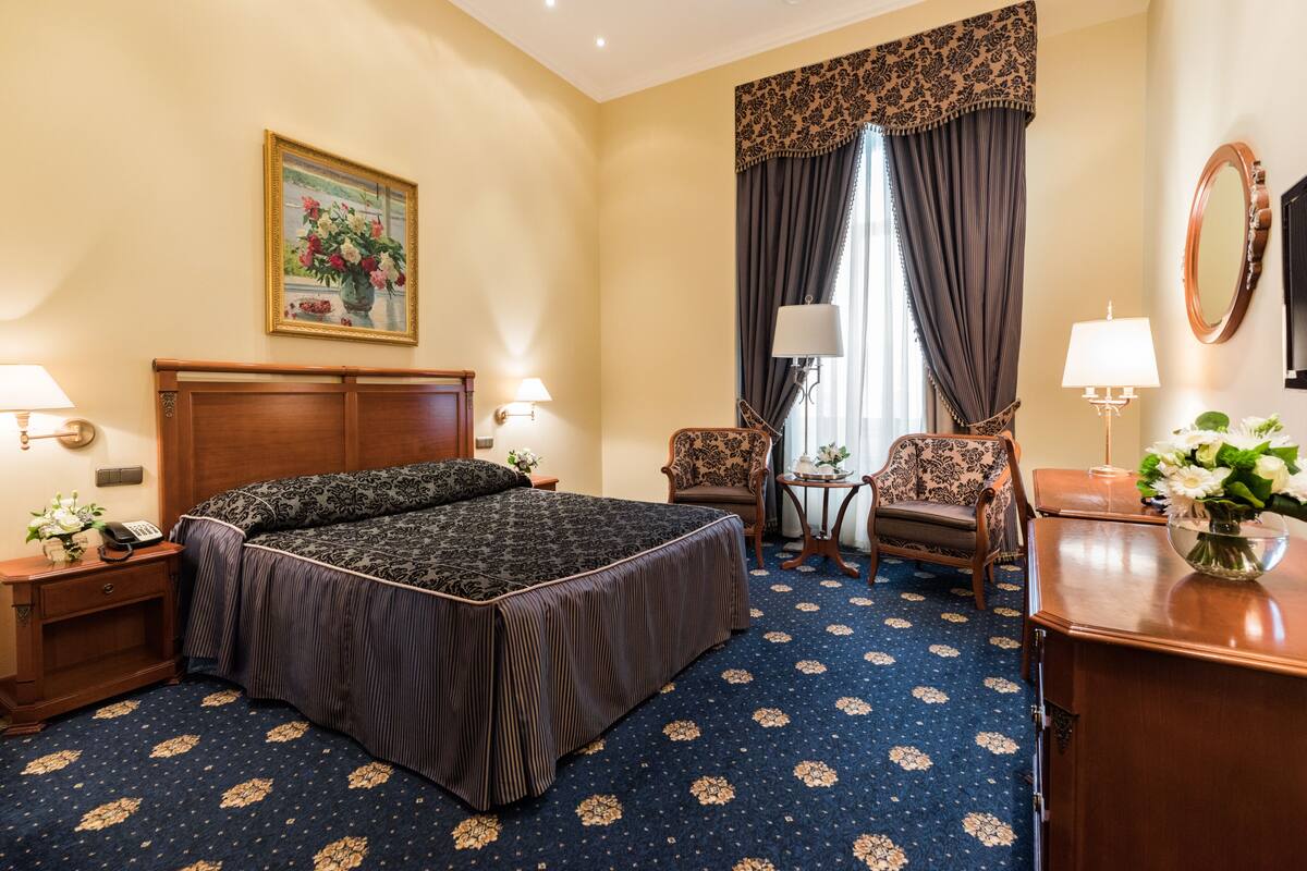 A hotel room with a large bed, two chairs, a desk, a lamp, and a painting on the wall.