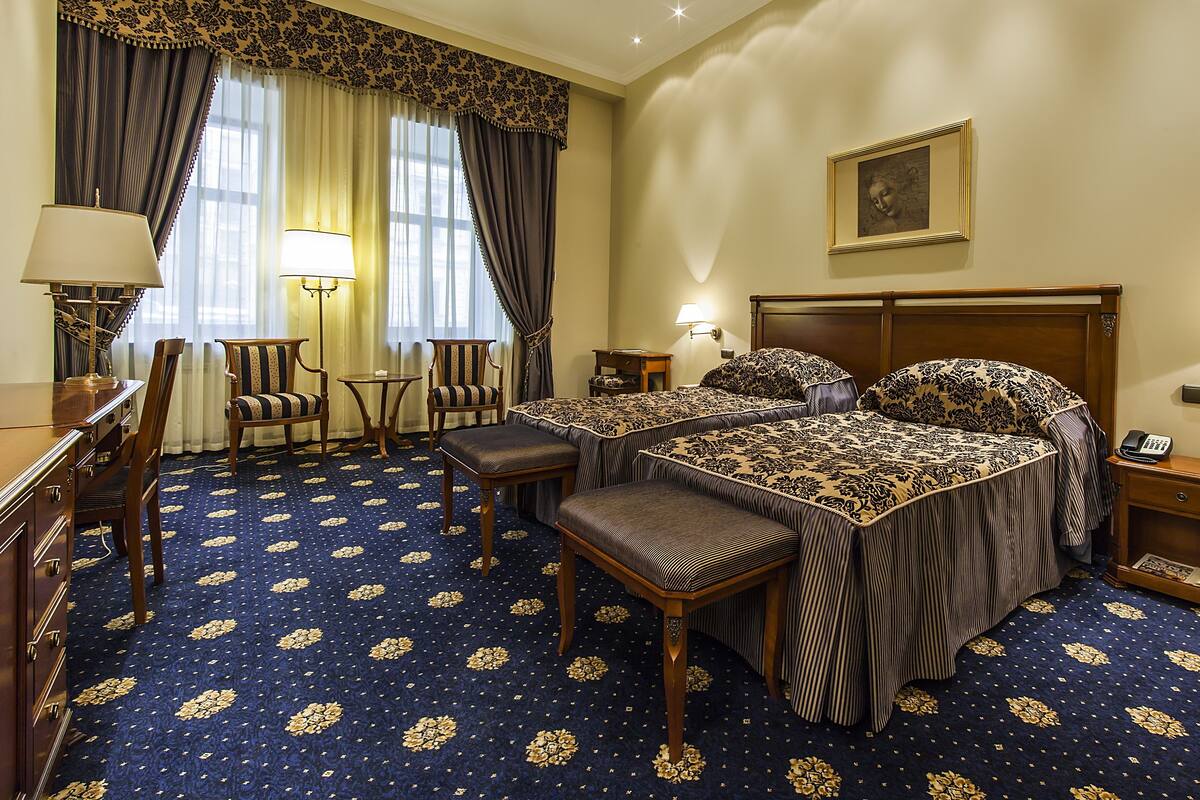 A hotel room with a large bed, two bedside tables, a desk with a chair, a lamp, and a patterned carpet.