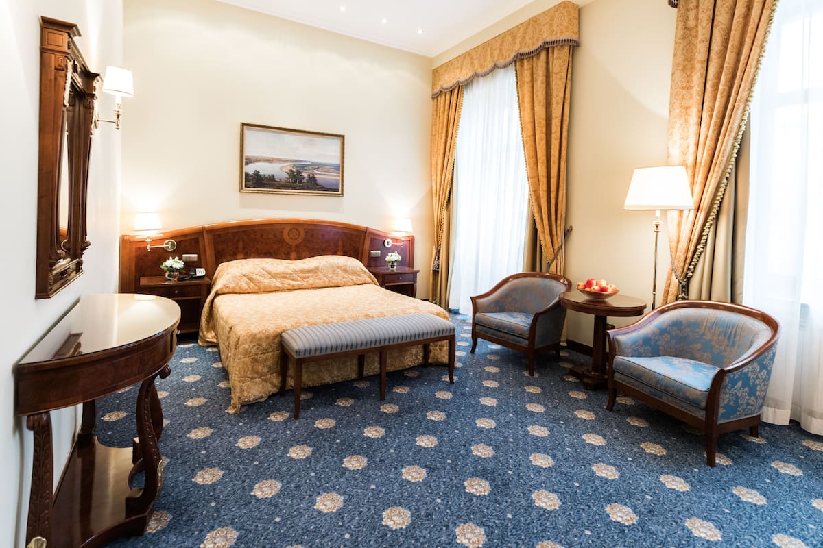 A hotel room with a large bed, a desk, two armchairs, a small table, and a lamp.