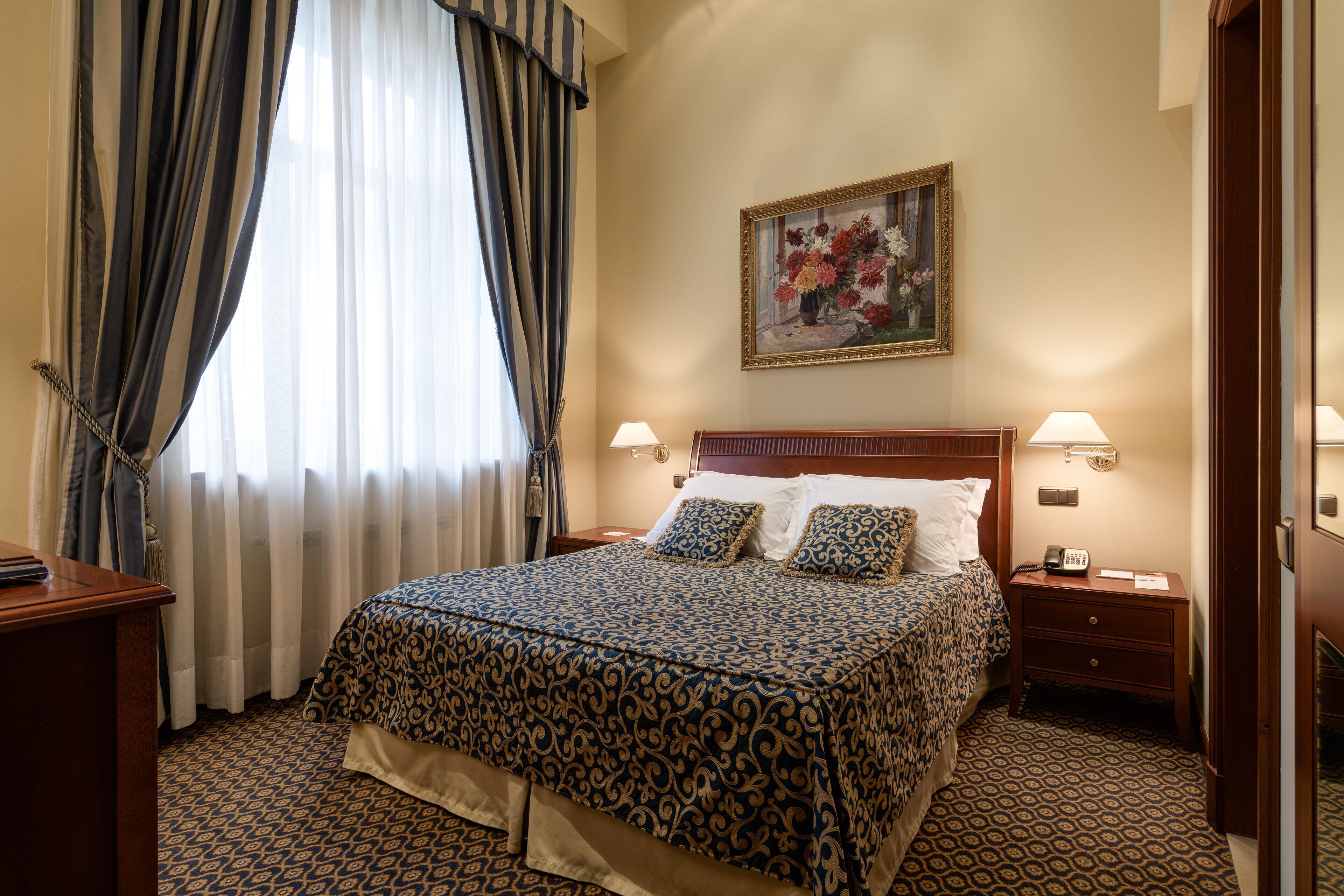 Habitación de hotel con una cama grande, dos mesitas de noche con lámparas, un cabecero de madera, un cuadro en la pared y una alfombra estampada.