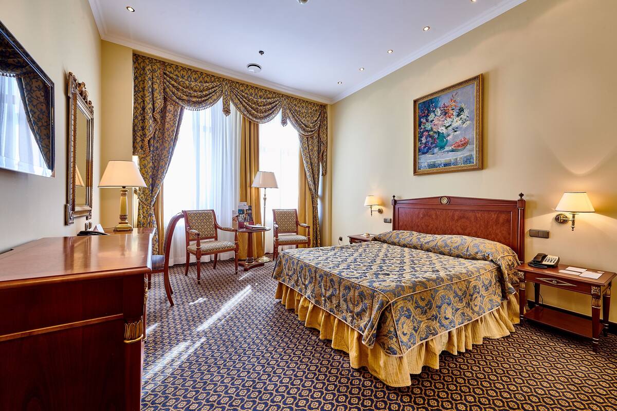 A hotel room with a large bed, two bedside tables with lamps, a desk with a chair, a large mirror, and a painting on the wall.