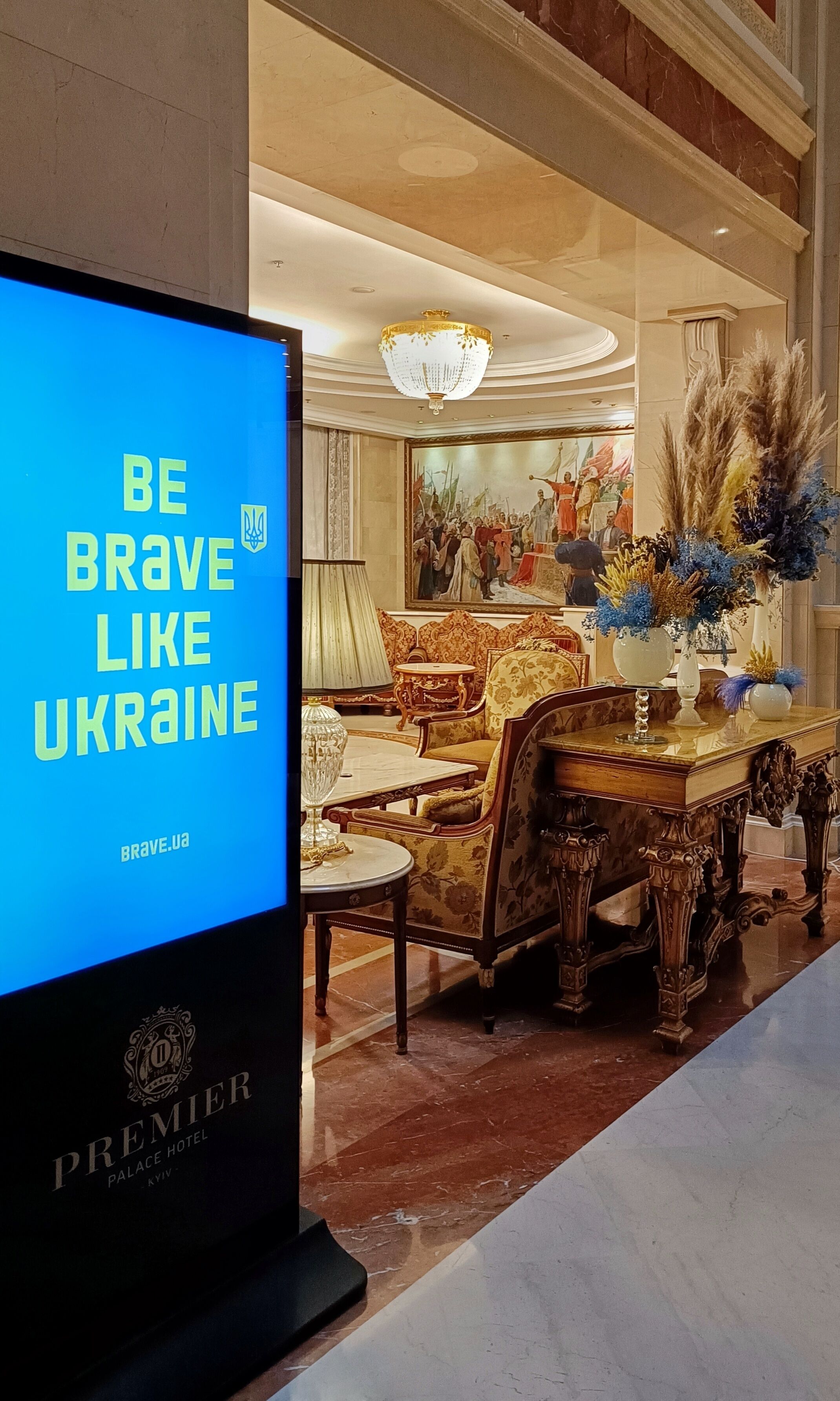 A hotel lobby with a large screen displaying "BE BRAVE LIKE UKRAINE" and a chandelier.