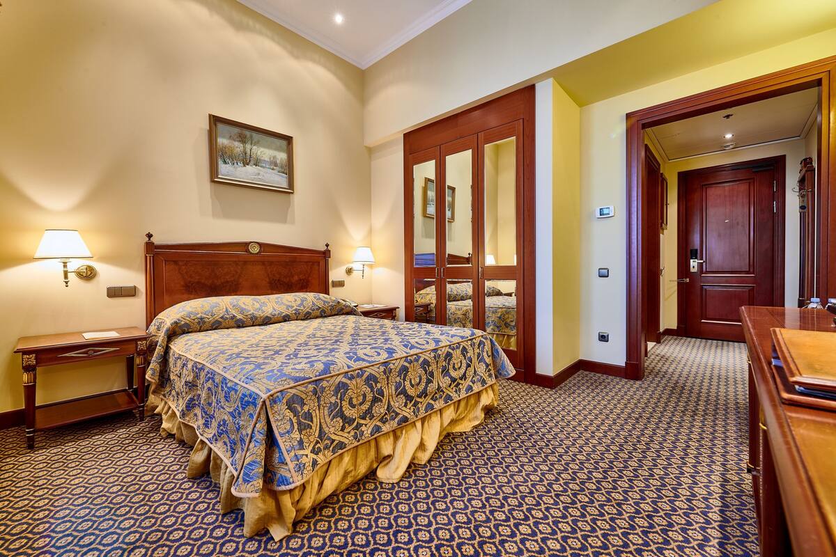 A hotel room with a large bed, two bedside tables, a nightstand, a wooden wardrobe, and a door leading to another room.