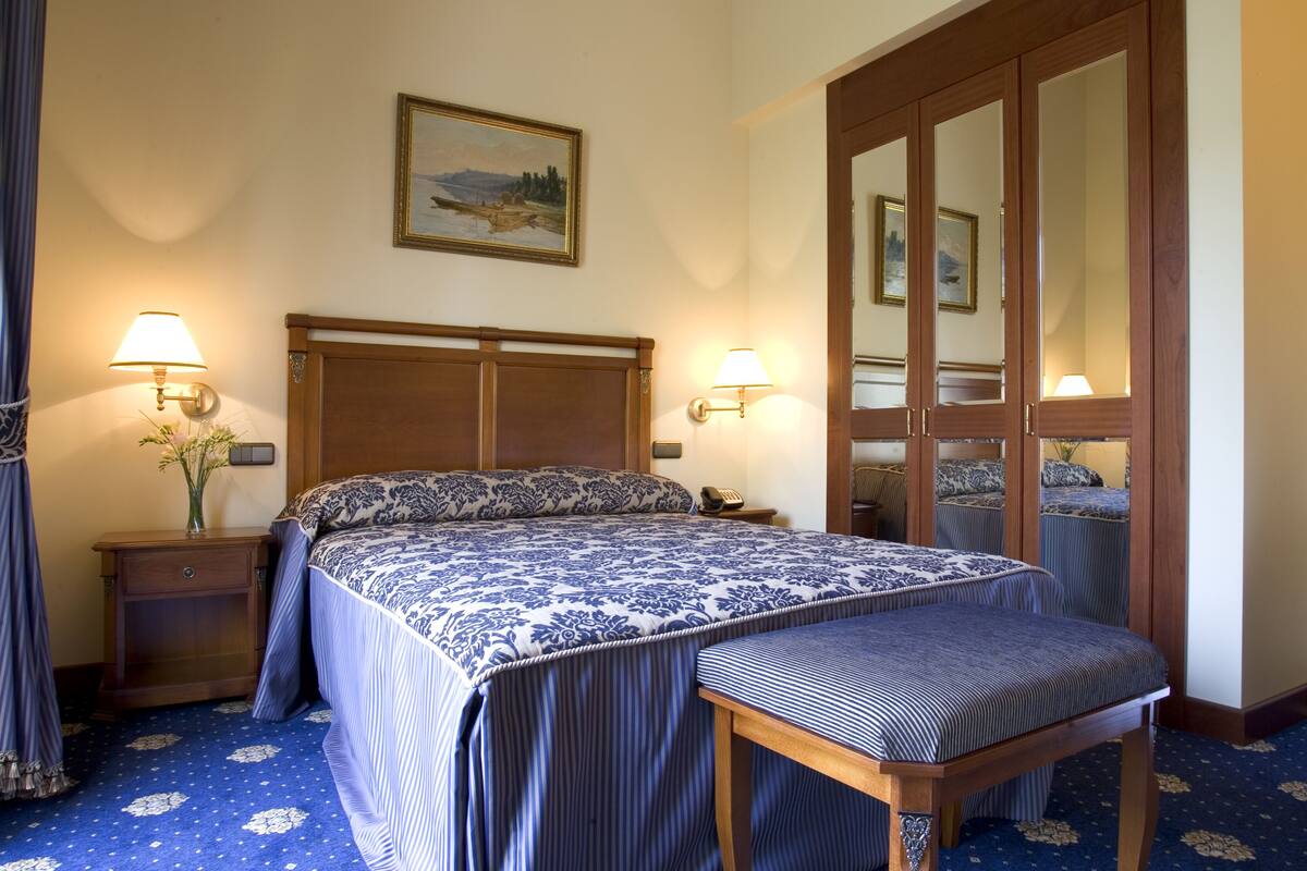 A hotel room with a large bed, two bedside lamps, a wooden nightstand, a bench, a mirror, and a painting on the wall.