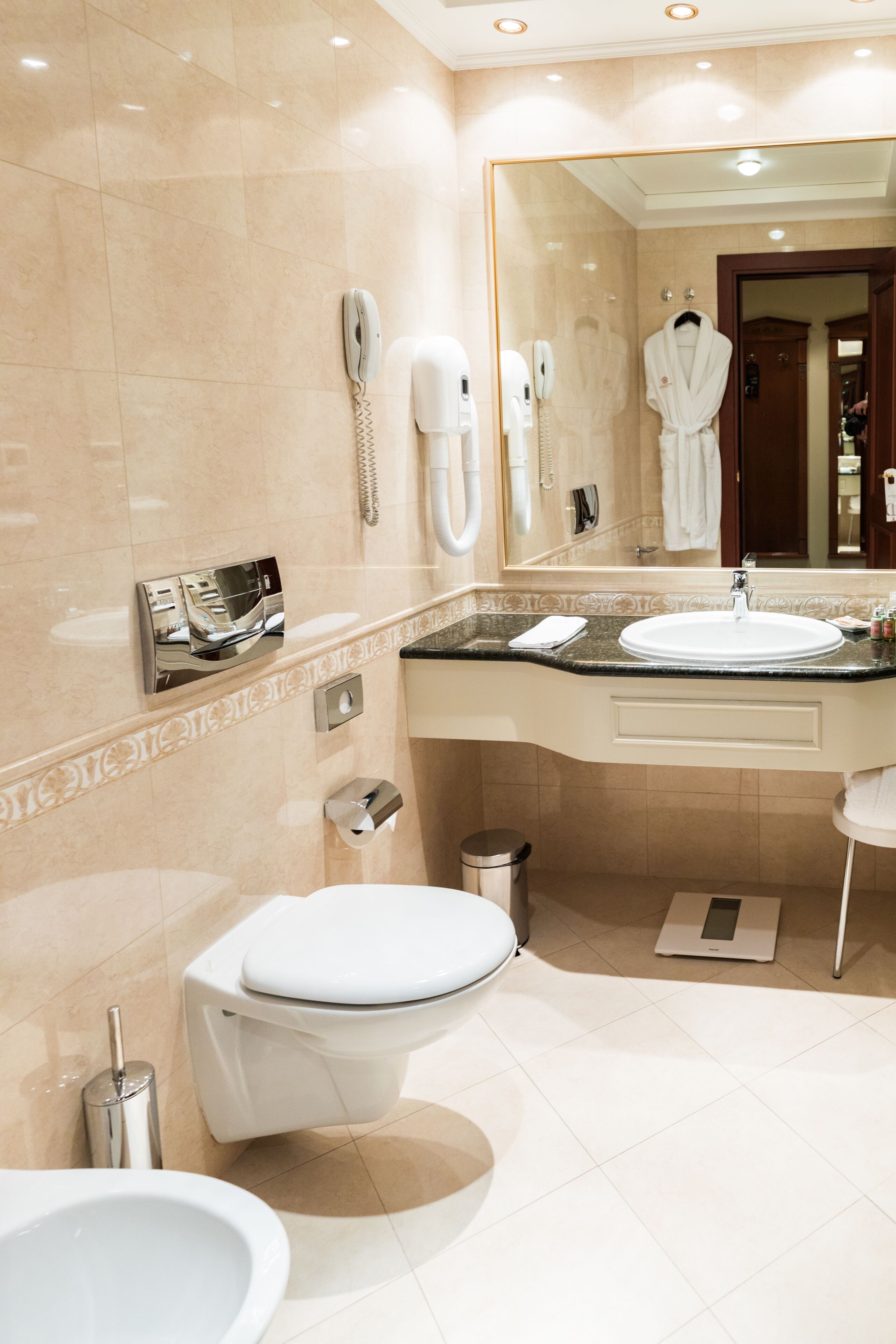 A modern bathroom with a white toilet, a sink, a bidet, a trash can, a hairdryer, and a mirror.