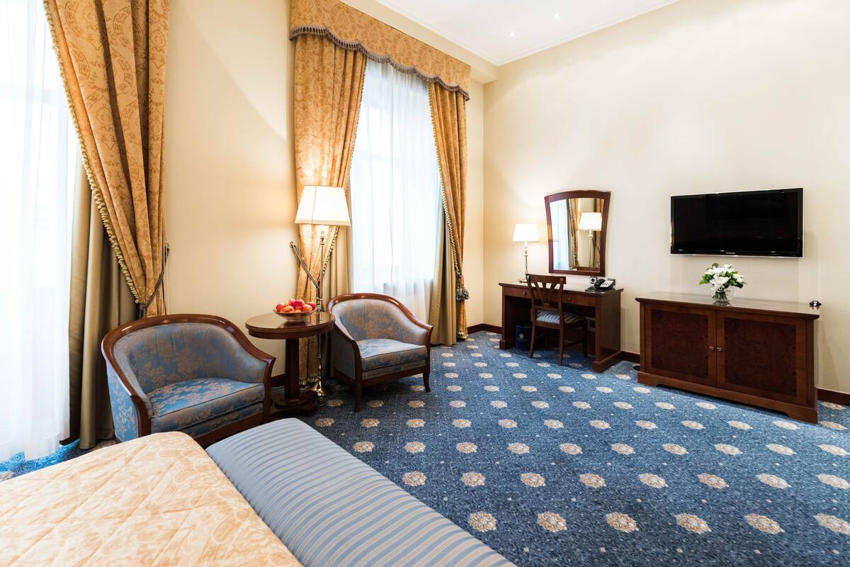 A hotel room with a large bed, two armchairs, a desk with a lamp, a television, and a patterned carpet.