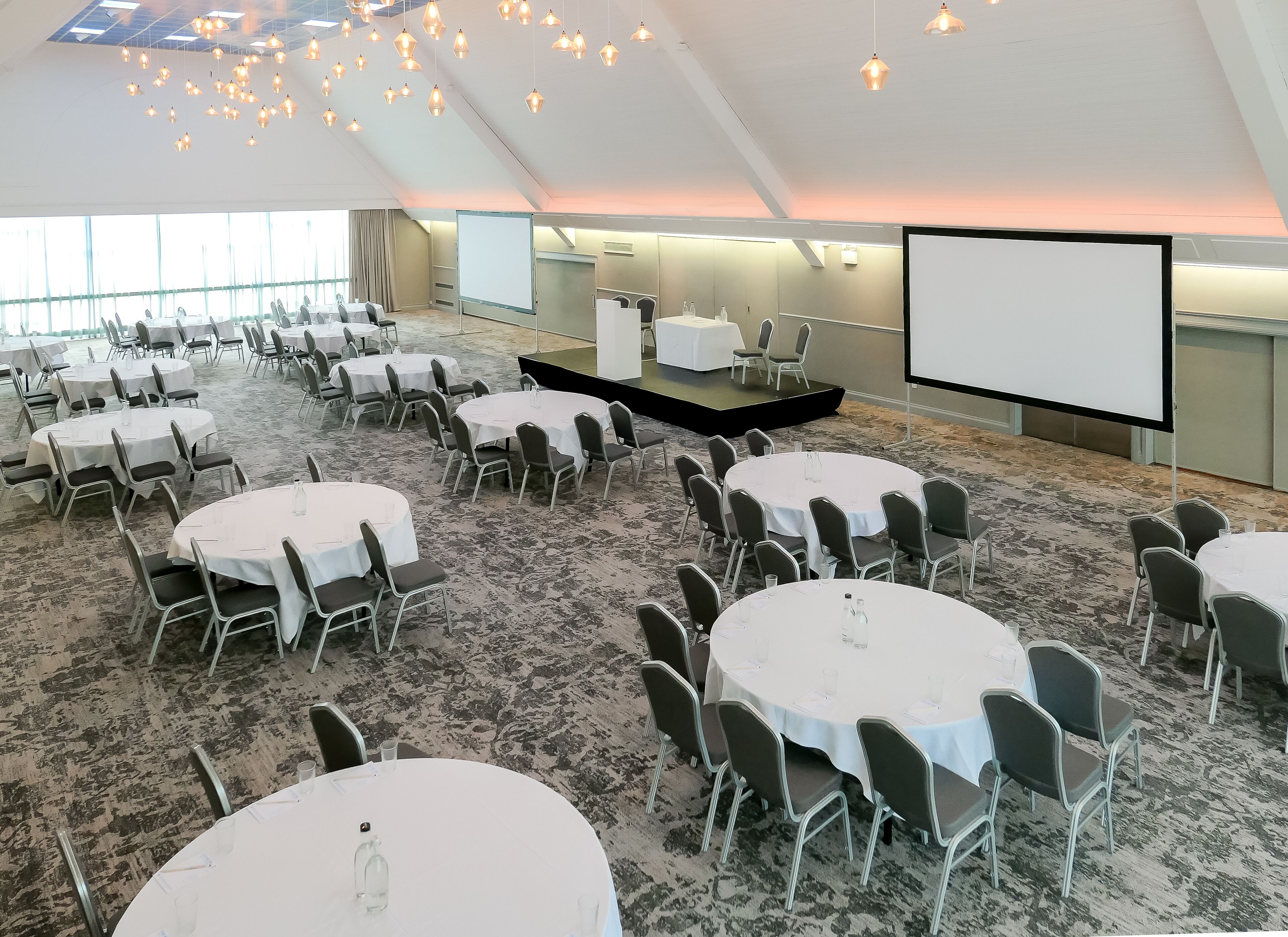 A conference room with multiple round tables and chairs, a stage with a screen, and a carpeted floor.