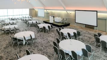 Un salón de conferencias con varias mesas redondas y sillas, un escenario con una pantalla, y suelo alfombrado.