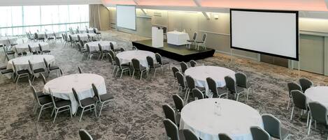 A conference room with multiple round tables and chairs, a stage with a screen, and a carpeted floor.