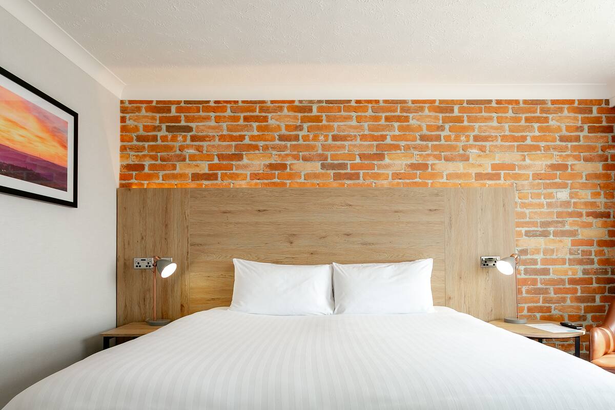 Habitación de hotel con pared de ladrillo, cabecero de madera, dos lámparas de noche, cama con ropa blanca y una silla de cuero marrón.