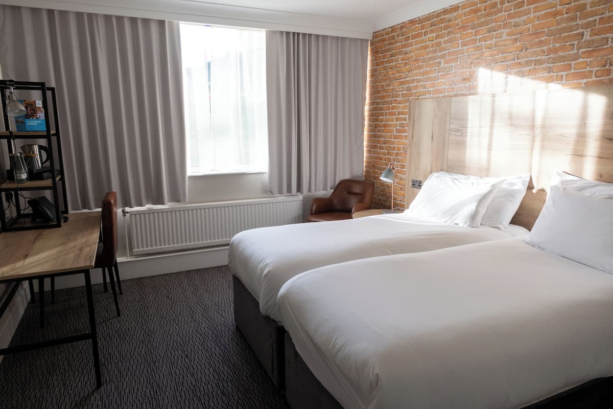 Habitación de hotel con dos camas, un escritorio de madera, una silla, un radiador y una pared de ladrillo.