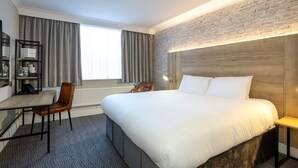 Habitación de hotel con una cama grande, un escritorio con silla, una estantería y una pared de ladrillo decorativa.