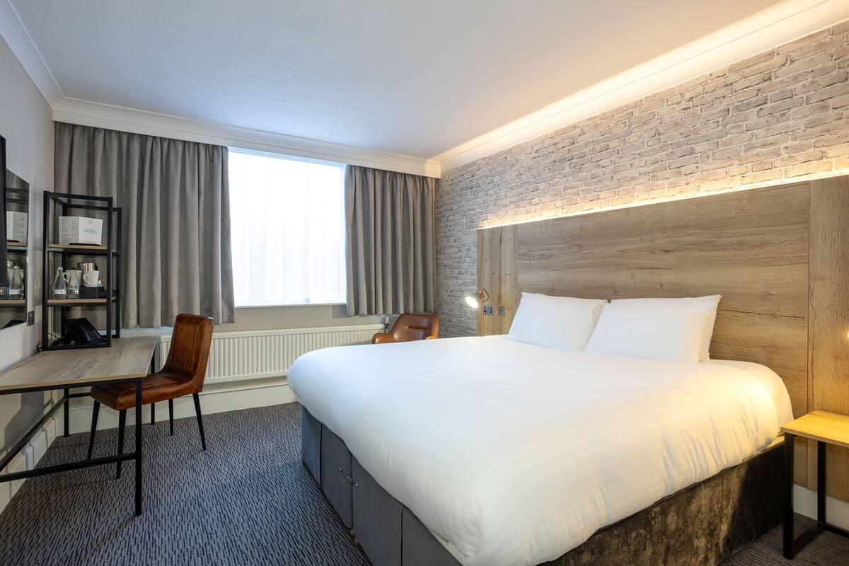 Habitación de hotel con una cama grande, un escritorio con silla, una estantería y una pared de ladrillo decorativa.
