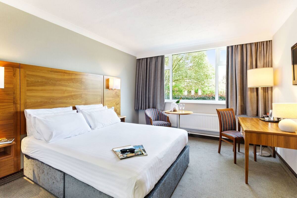 Una habitación de hotel con una cama grande, un escritorio, una silla, una ventana con cortinas y una lámpara.