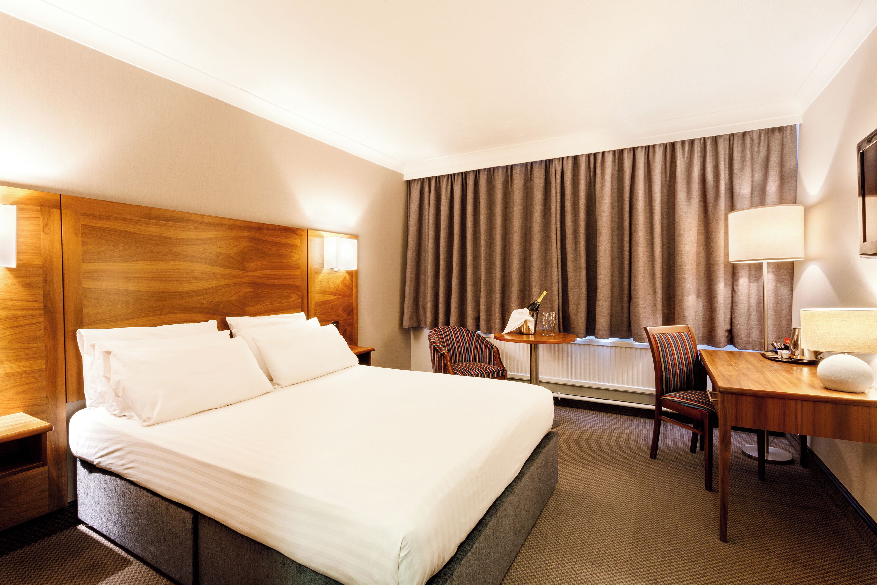 A hotel room with a large bed, two chairs, a desk with a lamp, and a view of the outdoors through curtains.
