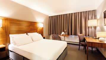 A hotel room with a large bed, two chairs, a desk with a lamp, and a view of the outdoors through curtains.