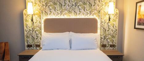 A hotel room with a large bed, two bedside lamps, a headboard with a leaf pattern, and a framed picture on the wall.