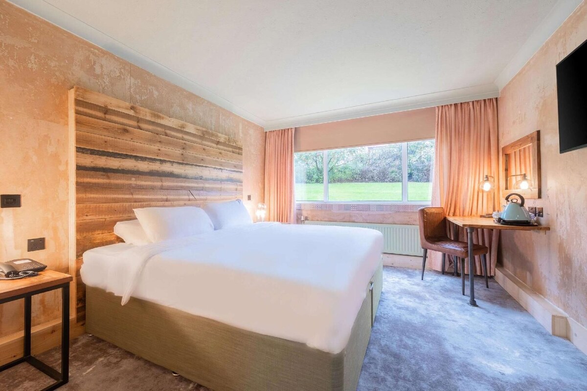 Hotelkamer met een groot bed, een bureau, een stoel, een telefoon en een raam met uitzicht op groen.