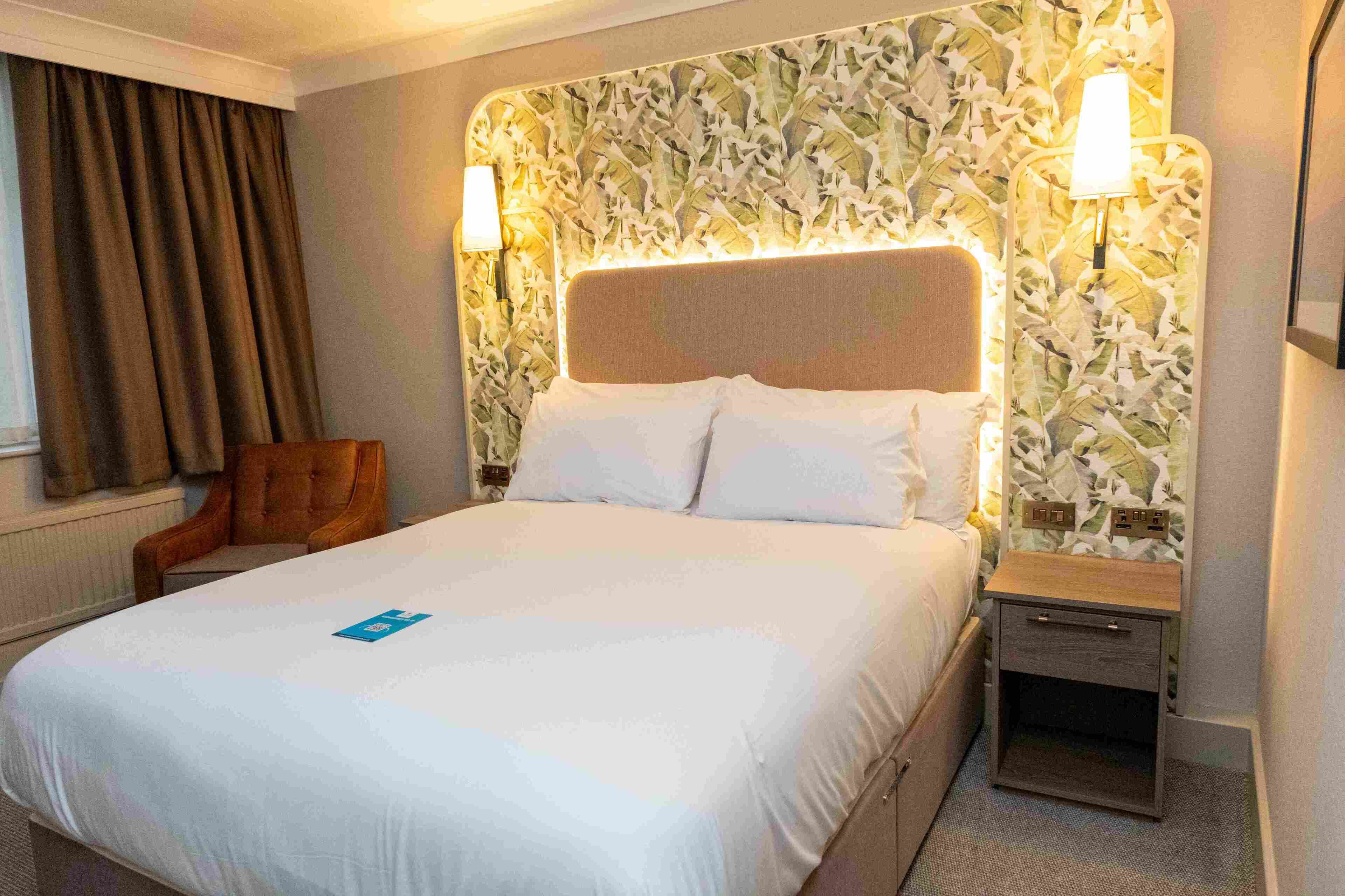 A hotel room with a large bed, two bedside tables, a chair, and a patterned headboard.