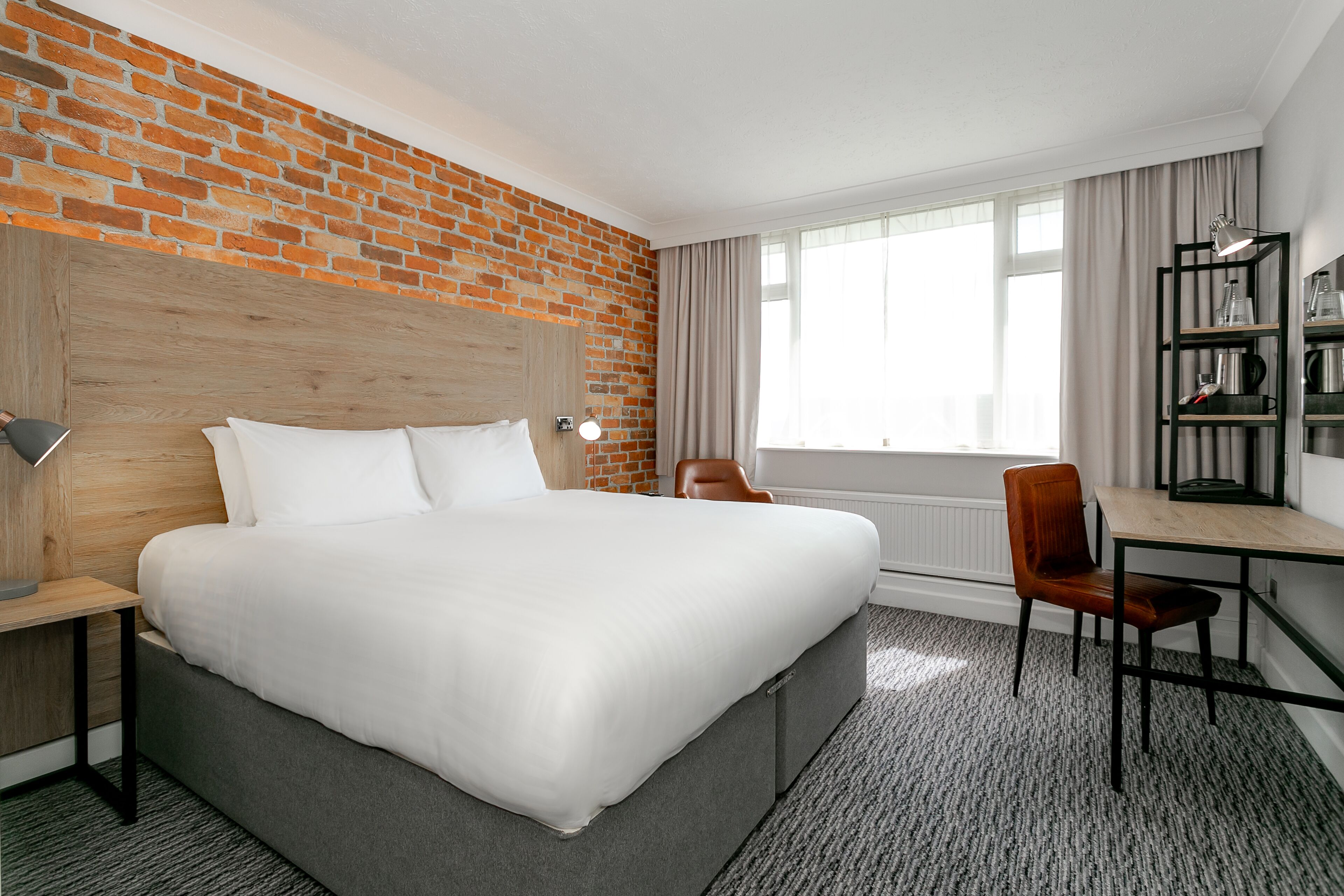 A hotel room with a brick wall, a large bed, a desk with a chair, and a bookshelf.