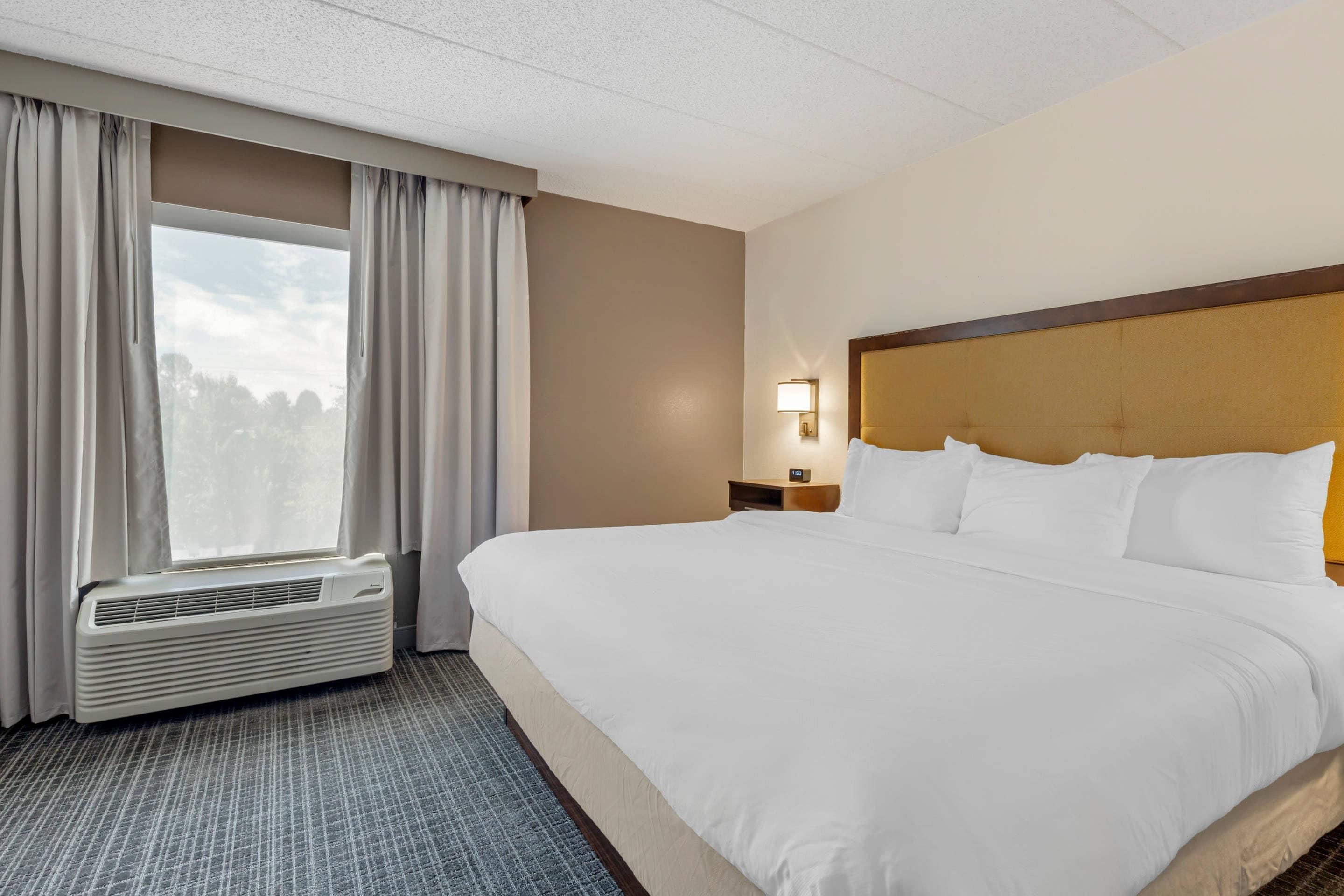 A hotel room with a large bed, a window with curtains, a wall-mounted light, and a thermostat.