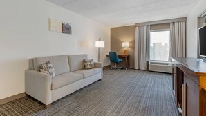 Habitación de hotel con sofá, escritorio, silla, lámpara, televisor y ventana con cortinas.