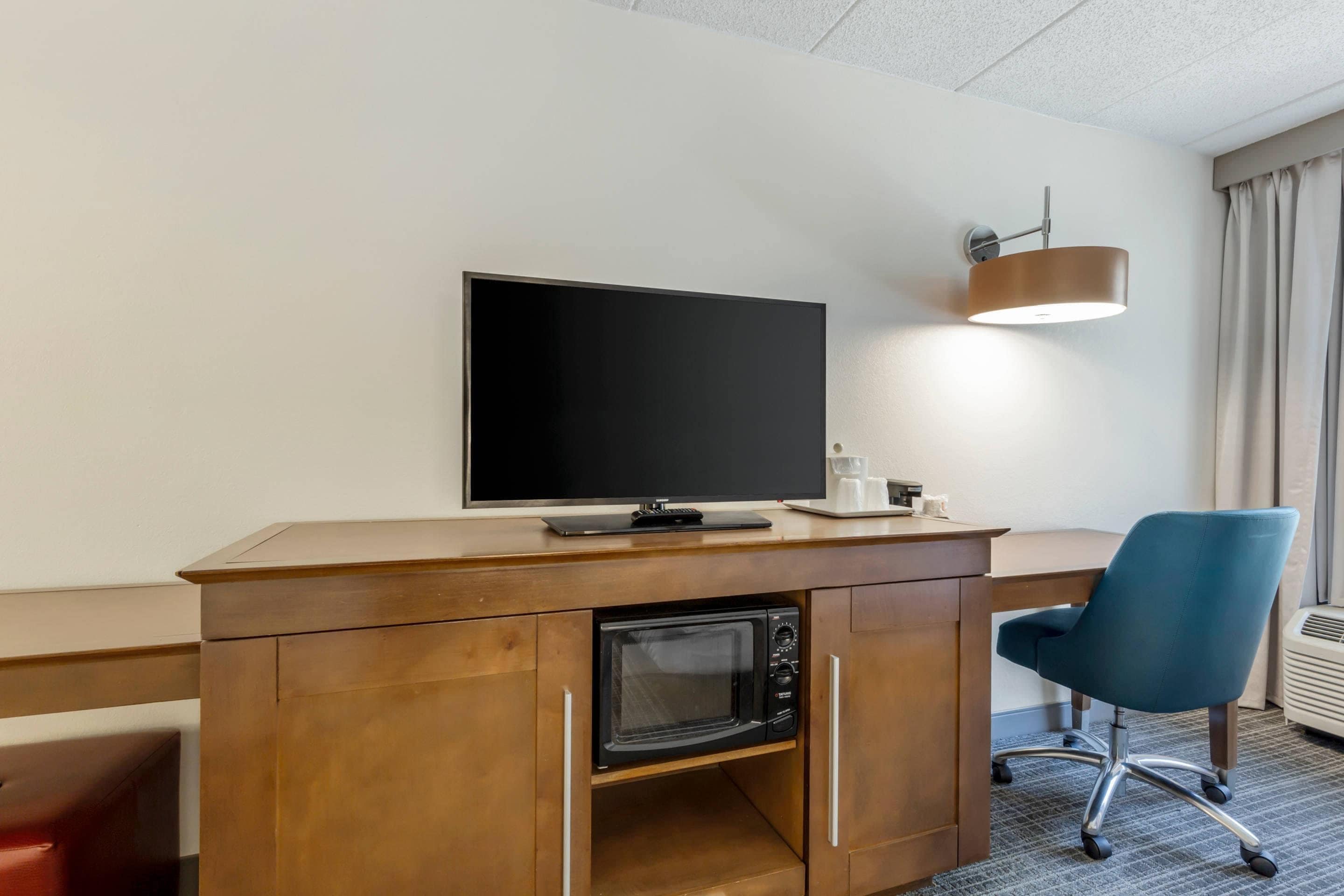 Habitación de hotel con un escritorio de madera, televisión, microondas, una silla de oficina azul y una lámpara de pared.