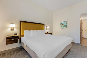 Habitación de hotel con una cama grande, dos mesitas de noche con lámparas, una mesita auxiliar y una obra de arte en la pared, además de una puerta.