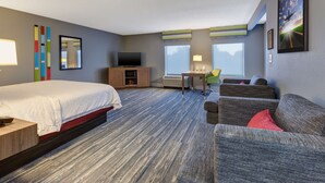 Quarto de hotel com cama, escrivaninha, televisão, sofá e uma janela grande.