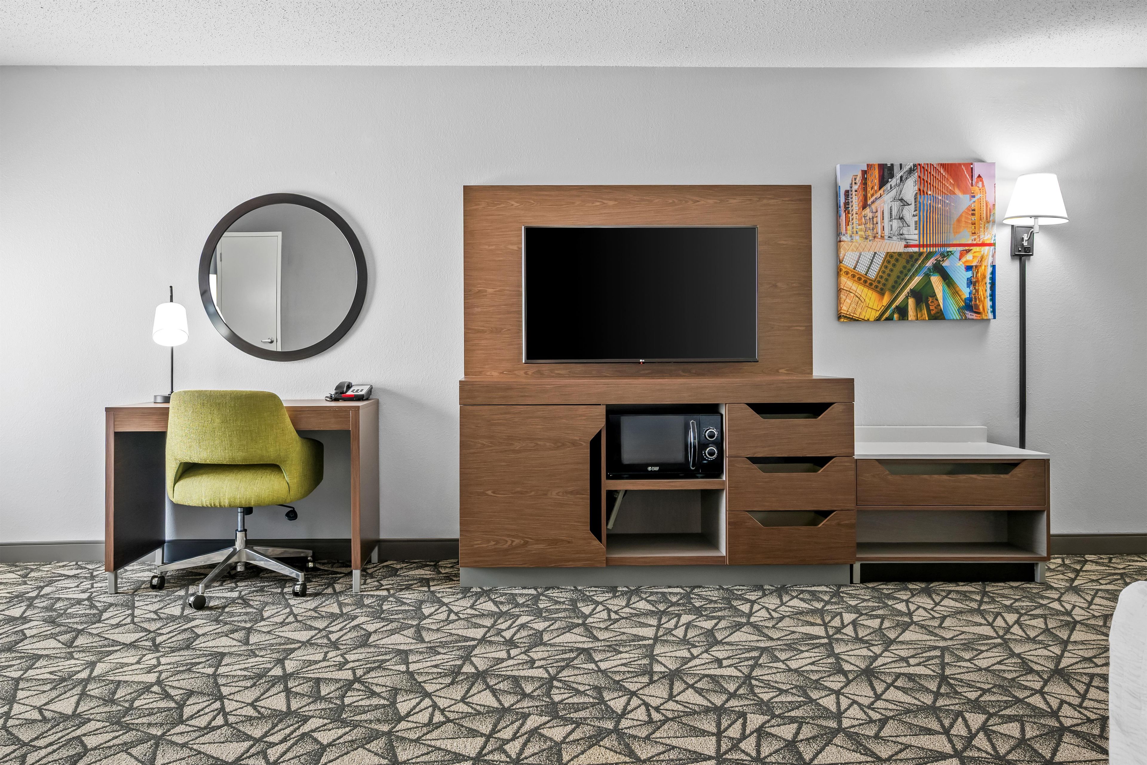 Habitación de hotel con televisión de pantalla plana, escritorio con lámpara, una silla verde, microondas y un cuadro en la pared.